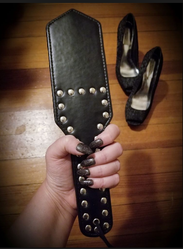 Ms. Sandalwood's hand holds a black studded paddle; a pair of black Vera Wang high-heeled shoes is visible in the background.